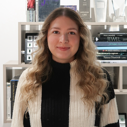 A woman with long wavy blonde hair wearing a black and cream striped ribbed knit sweater, standing in front of a white bookshelf filled with books and awards.